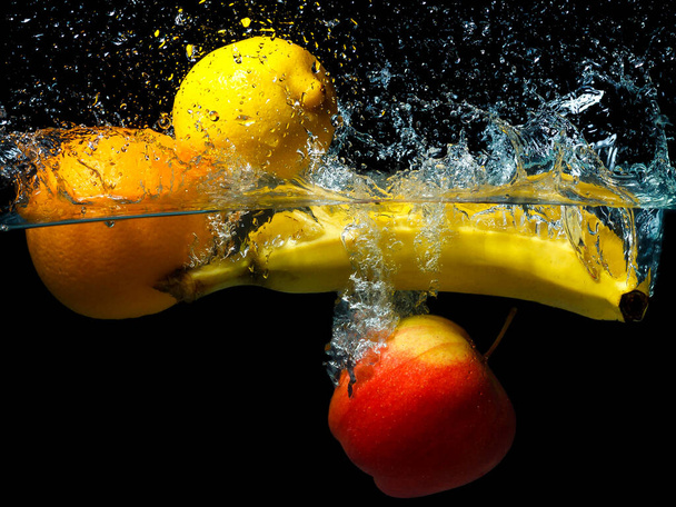 whole banana, orange, lemon and apple in water on a black background. Fresh fruit with water spray. - Фото, изображение
