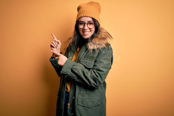 Giovane donna bruna indossa occhiali e cappotto invernale con cappello su sfondo giallo isolato sorridente e guardando la fotocamera che punta con due mani e dita di lato
. - Foto, immagini
