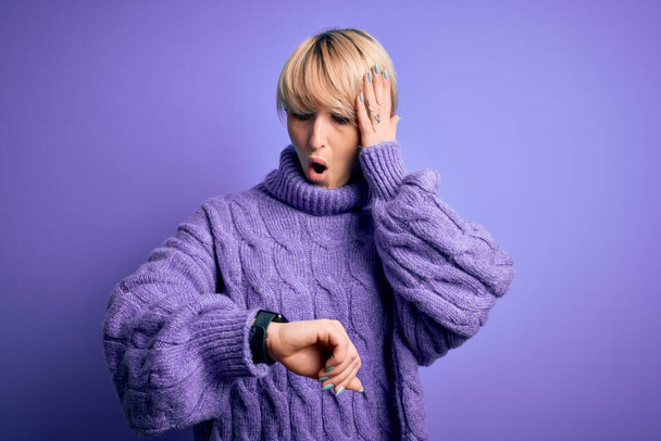 Молода блондинка з коротким волоссям, одягнена в зимовий светр черепашки на фіолетовому фоні Дивлячись, як годинник турбується, боїться запізнюватися
 - Фото, зображення