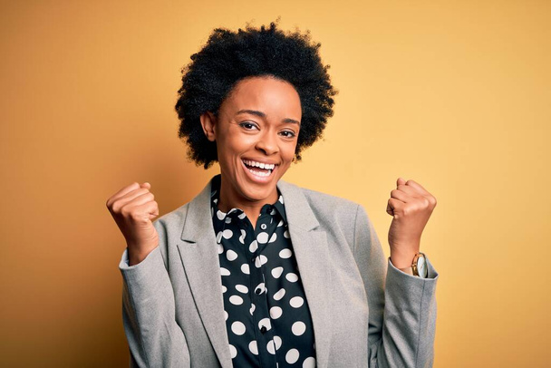 Jonge mooie Afro-Amerikaanse afro zakenvrouw met krullend haar dragen jas vieren verrast en verbaasd voor succes met armen opgeheven en open ogen. Winnaars concept. - Foto, afbeelding