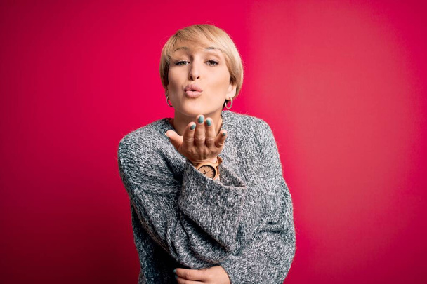 Young blonde woman with modern short hair wearing casual sweater over pink background looking at the camera blowing a kiss with hand on air being lovely and sexy. Love expression. - 写真・画像