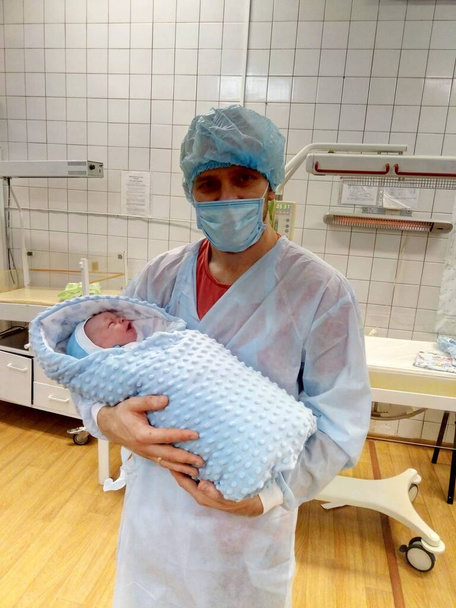 Muž světlé pleti v jednorázovém ochranném oděvu drží novorozeně na porodním sále. - Fotografie, Obrázek