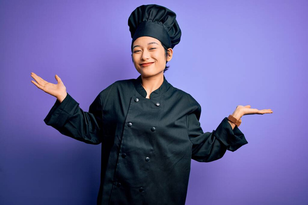 Nuori kaunis kiinalainen kokki nainen yllään liesi yhtenäinen ja hattu yli violetti tausta tietämätön ja sekava ilme kädet ja kädet nostetaan. Epävarma käsite
. - Valokuva, kuva