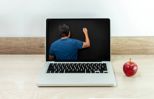 Laptop op een bureau met een appel ernaast en een leraar op zijn rug die op een schoolbord op zijn monitor schrijft - Foto, afbeelding