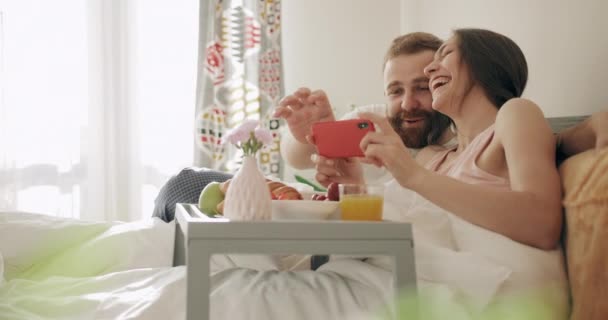 Cheerful family watching photos while sitting on bed with tray full of food. Young woman and man having breakfast in bed, laughing while looking and touching smartphone screen. - Séquence, vidéo