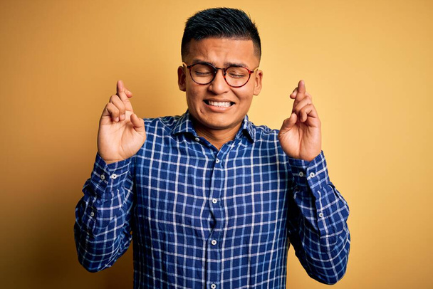 Young handsome latin man wearing casual shirt and glasses over yellow background gesturing finger crossed smiling with hope and eyes closed. Luck and superstitious concept. - Foto, imagen