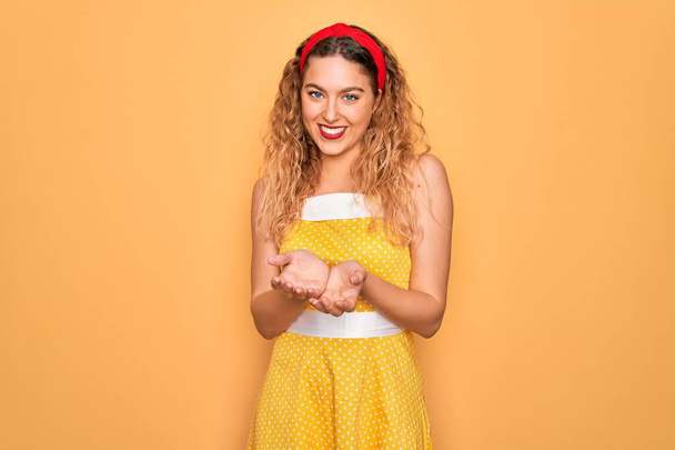 Beautiful blonde pin-up woman with blue eyes wearing diadem standing over yellow background Smiling with hands palms together receiving or giving gesture. Hold and protection - Fotografie, Obrázek