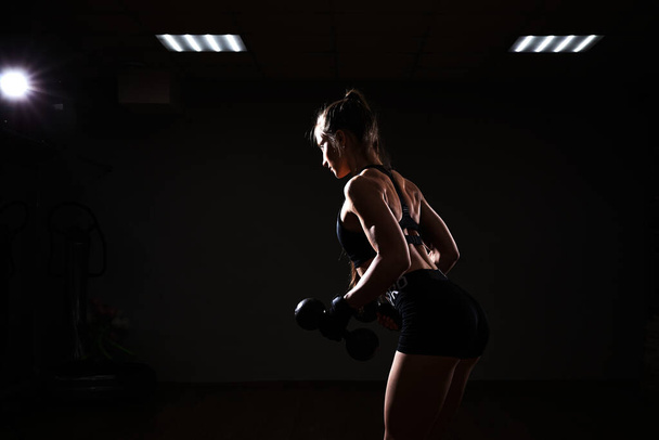 フィットネスとスポーツのコンセプト。残酷な運動女性ポンプアップmusclesとともにダンベル上の暗い背景. - 写真・画像