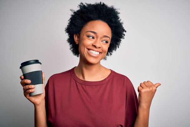 Fiatal afro-amerikai nő göndör haj ivás csésze kávét mutat, és mutatja hüvelykujjával oldalra boldog arc mosolygós - Fotó, kép