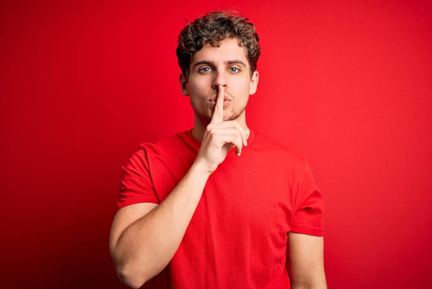 Ein junger blonder, gutaussehender Mann mit lockigem Haar trägt ein lässiges T-Shirt über rotem Hintergrund und bittet mit dem Finger auf den Lippen um Ruhe. Schweigen und geheimes Konzept. - Foto, Bild