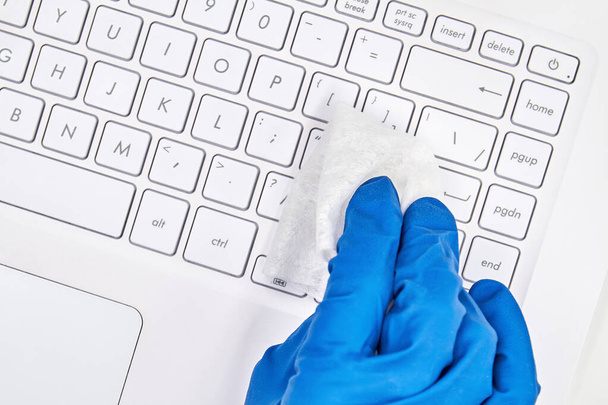 Ręka kobiety w niebieskiej rękawicy ochronnej czyści się środkiem dezynfekującym powierzchnię laptopa do zapobiegania koronawirusowi, bakteriom, wirusom i zarazkom. Zbliżenie, selektywne ukierunkowanie - Zdjęcie, obraz