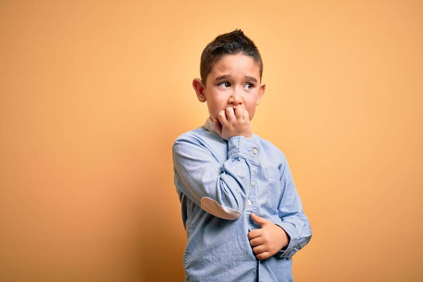 Маленький мальчик в элегантной рубашке, стоящей на желтом изолированном фоне, выглядящий напряженным и нервным с руками на рту, грызущими ногти. Проблема тревоги
. - Фото, изображение