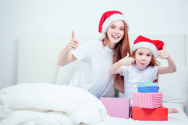 beautiful redhaired ginger woman with santa claus hat and happy little girl in pajamas sitting on bed showing thumbs up gesture next to gifts boxes.christmas morning celebration - Foto, Bild