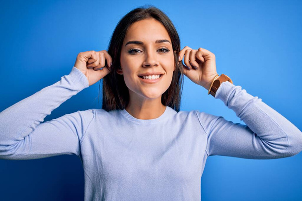Young beautiful brunette woman wearing casual sweater standing over blue background Smiling pulling ears with fingers, funny gesture. Audition problem - Photo, Image