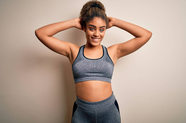 Young african american sportswoman doing sport wearing sportswear over white background relaxing and stretching, arms and hands behind head and neck smiling happy - Photo, image
