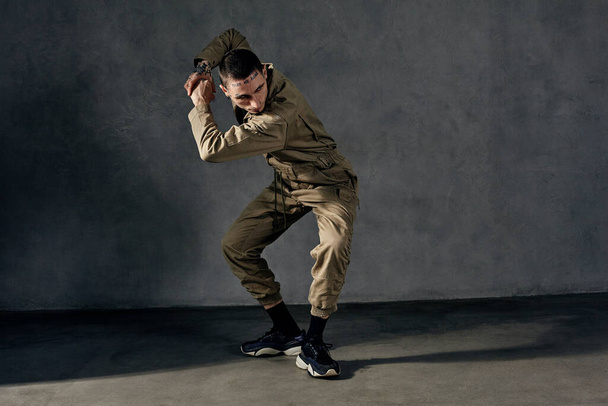 Young graceful guy with tattooed body and face, earrings, beard. Dressed in khaki overalls and black sneakers. He is dancing against gray studio background. Dancehall, hip-hop. Full length, copy space - 写真・画像