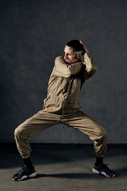Young graceful performer with tattooed body and face, earrings, beard. Dressed in khaki overalls and black sneakers. He is dancing against gray background. Dancehall, hip-hop. Full length, copy space - Photo, Image