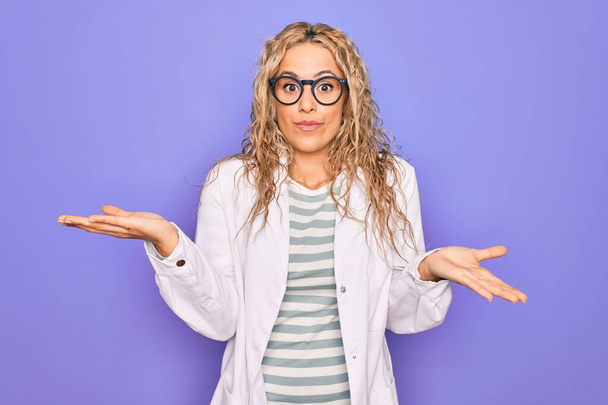 Young beautiful blonde scientist woman wearing coat and glasses over purple background clueless and confused expression with arms and hands raised. Doubt concept. - Photo, Image