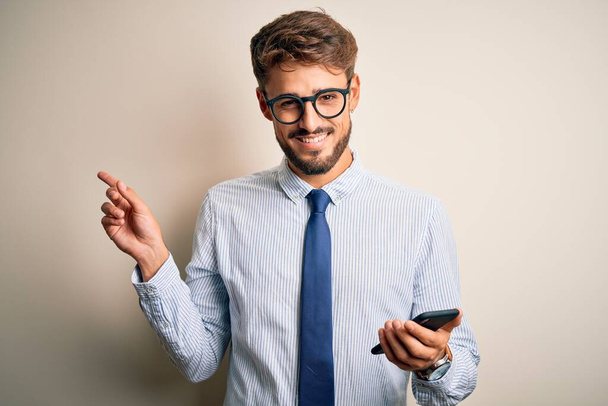 Jonge zakenman die een gesprek met behulp van smartphone over witte achtergrond zeer gelukkig wijzen met de hand en vinger naar de zijkant - Foto, afbeelding