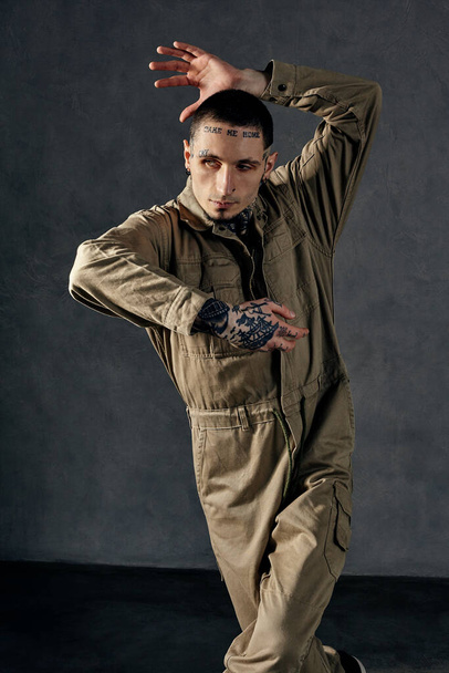 Young modern male with tattooed body and face, earrings, beard. Dressed in khaki jumpsuit and black sneakers. He is dancing against gray studio background. Dancehall, hip-hop. Full length, copy space - Foto, immagini