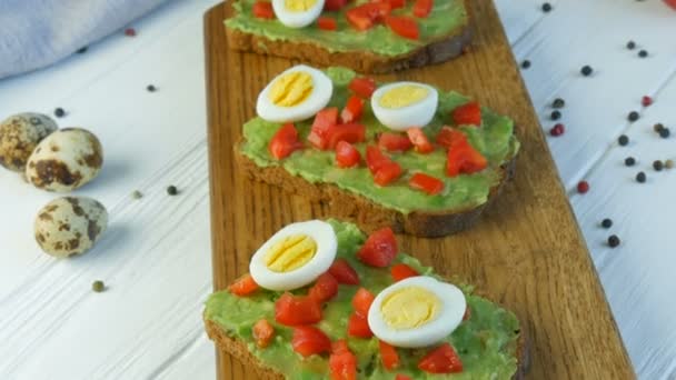 Healthy Vegan food. Spread mashed avocado on a toasted brown bread. Making tasty avocado toast for breakfast. Cooking bruschetta with cherry tomatoes and quail eggs. - Footage, Video