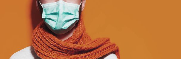 Young guy with scarf, wearing medical respiratory face mask against coronavirus. Panoramic portrait on light brown background with copy space. - Photo, Image