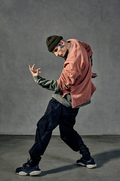 Young handsome guy with tattooed body and face, earrings, beard. Dressed in hat, casual clothes and black sneakers. Dancing on gray background. Dancehall, hip-hop. Full length, copy space - 写真・画像