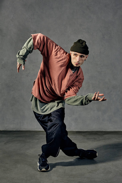 Young handsome performer with tattooed body and face, earrings, beard. Dressed in hat, casual clothes and black sneakers. Dancing on gray background. Dancehall, hip-hop. Full length, copy space - Фото, зображення