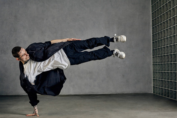 Flexible guy with tattooed body, earrings, beard. Dressed in white t-shirt and sneakers, black denim shirt and pants. Showing tricks on gray background. Dancehall, hip-hop. Full length, copy space - Photo, Image