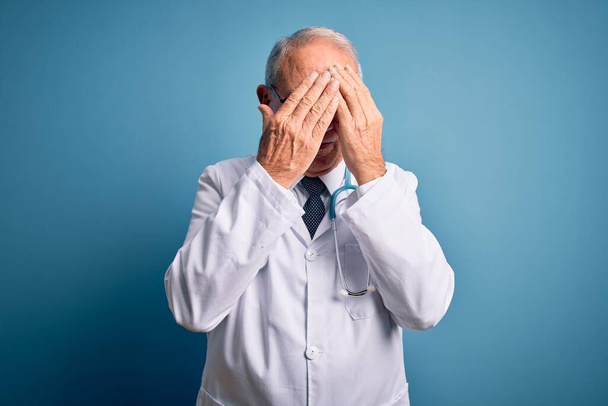 Hombre médico de pelo gris senior que usa estetoscopio y abrigo médico sobre fondo azul con expresión triste que cubre la cara con las manos mientras llora. Concepto de depresión
. - Foto, imagen