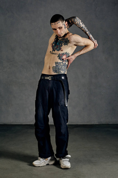 Flexible male with tattooed body and face, naked torso, beard. Dressed in black pants and white sneakers. He is dancing on gray studio background. Dancehall, hip-hop. Full length, copy space - 写真・画像