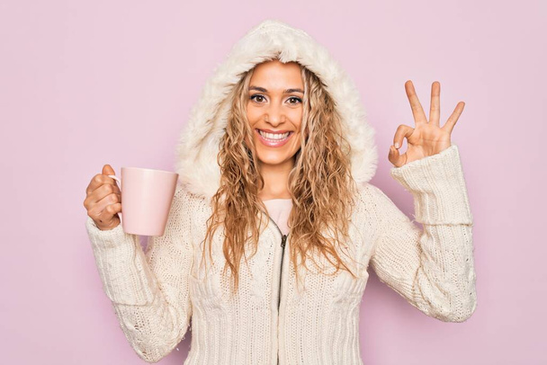 Fiatal gyönyörű szőke nő visel alkalmi pulóver motorháztető ivás csésze kávét csinál rendben jel ujjakkal, mosolygós barátságos gesztus kiváló szimbólum - Fotó, kép
