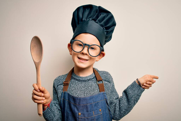Young little caucasian cook kid wearing chef uniform and hat cooking using wooden spoon very happy pointing with hand and finger to the side - Photo, Image