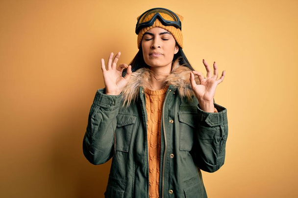 Νεαρή όμορφη ισπανόφωνη γυναίκα που φοράει γυαλιά σκι και παλτό για χειμερινό καιρό χαλαρό και χαμογελαστό με τα μάτια κλειστά κάνει χειρονομία διαλογισμού με τα δάχτυλα. Έννοια γιόγκα. - Φωτογραφία, εικόνα
