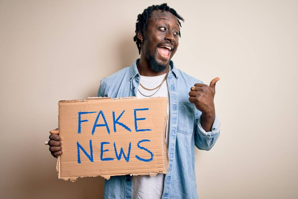 Jovem afro-americano segurando banner com mensagem de notícias falsas sobre fundo branco apontando e mostrando com o polegar para o lado com o rosto feliz sorrindo
 - Foto, Imagem