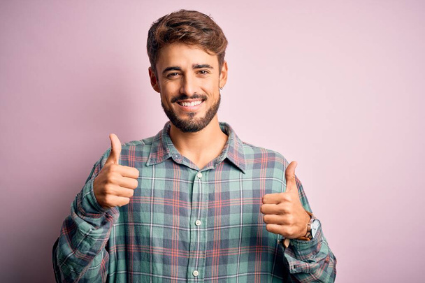 Mladý pohledný muž s vousy v neformální košili stojící nad růžovým pozadím znamení úspěchu dělá pozitivní gesto rukou, palce nahoru s úsměvem a šťastný. Veselý výraz a gesto vítěze. - Fotografie, Obrázek
