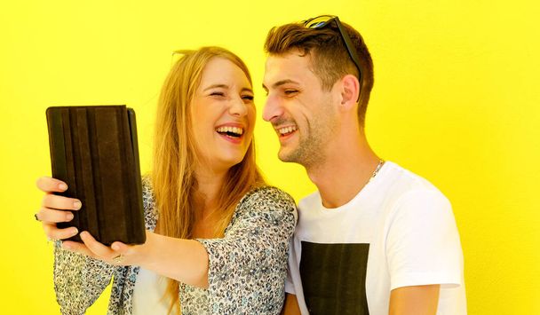 Feliz pareja joven usando tableta digital y teléfono celular fuera en la temporada de verano en el fondo amarillo de la ciudad
 - Foto, Imagen