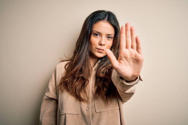 Young beautiful brunette woman wearing casual shirt standing over white background doing stop sing with palm of the hand. Warning expression with negative and serious gesture on the face. - 写真・画像