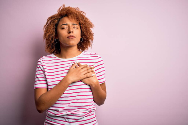 Young beautiful African American afro woman with curly hair wearing casual striped t-shirt smiling with hands on chest with closed eyes and grateful gesture on face. Health concept. - Photo, Image