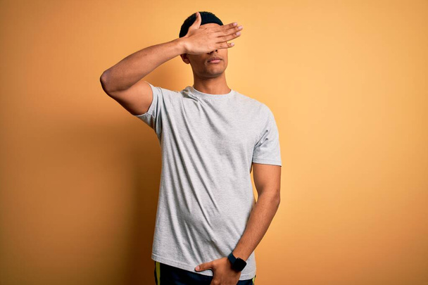 Handsome african american sportsman doing sport wearing sportswear over yellow background covering eyes with hand, looking serious and sad. Sightless, hiding and rejection concept - Photo, image