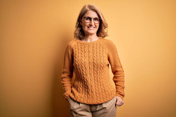 Middle age beautiful blonde woman wearing casual sweater and glasses over yellow background with a happy and cool smile on face. Lucky person. - Photo, Image