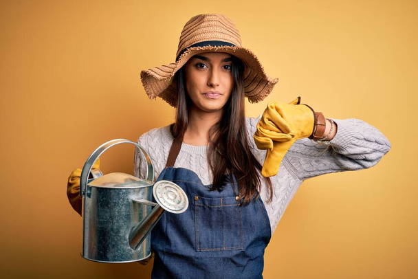 若い美しいブルネット農家の女性を身に着けているエプロンと帽子を保持する散水缶とともに怒っている顔,否定的な兆候ショー嫌いで親指ダウン,拒絶概念 - 写真・画像
