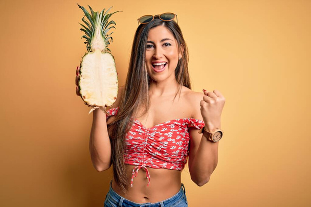 Young beautiful brunette woman holding middle pineapple fruit over isolated yellow background screaming proud and celebrating victory and success very excited, cheering emotion - Fotó, kép