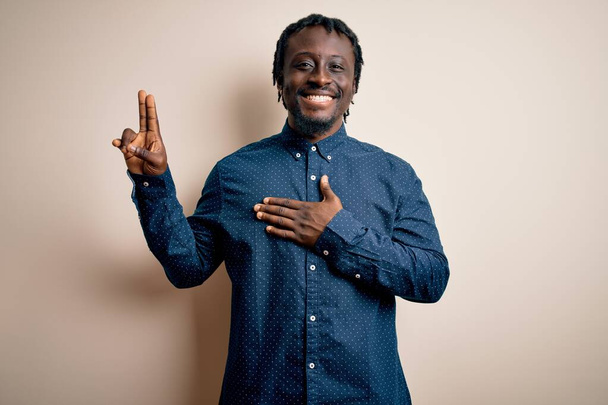 Mladý pohledný africký Američan muž v ležérní košili stojící nad bílým pozadím s úsměvem nadávky s rukou na hrudi a prsty nahoru, věrnostní slib přísahu - Fotografie, Obrázek