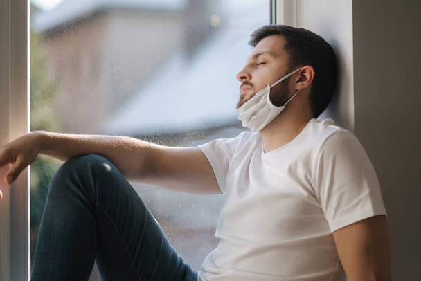 Young man sits on windowsill and pulled protective mask below on the chin and look s out the window. Alone at home in quarantine. Coronavirus theme. Stay at home - Foto, Bild
