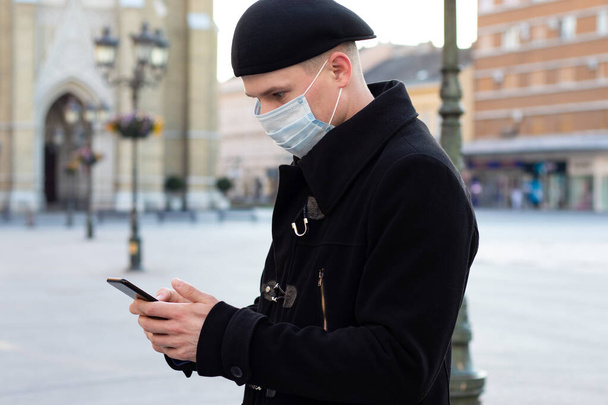 Junger Mann mit Schutzmaske im Gesicht steht allein auf der leeren Straße und wartet auf ein Date, während er auf seinem Smartphone nach Daten sucht. Covid 19 Virus-Pandemie oder Luftverschmutzungskonzept. - Foto, Bild