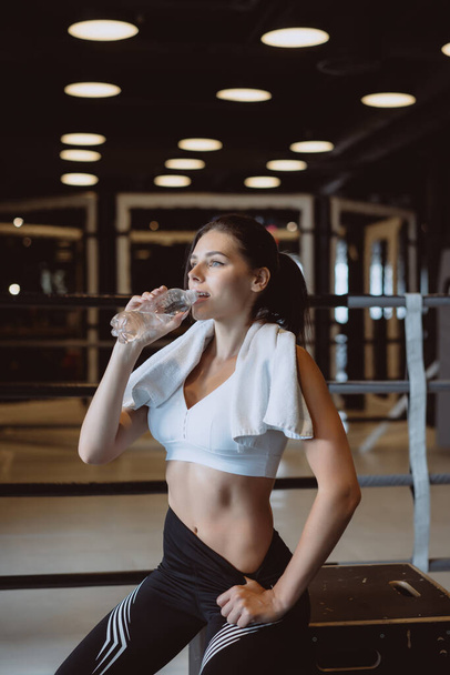 Hermosa joven con una toalla en los hombros bebiendo agua de una botella en el gimnasio
 - Foto, Imagen