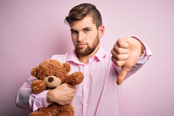 Young blond man with beard and blue eyes holding teddy bear toy over pink background with angry face, negative sign showing dislike with thumbs down, rejection concept - 写真・画像