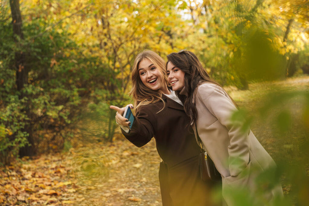 Two cheerful young pretty girls wearing coats walking together at the autumn forest, pointing finger away - Фото, изображение
