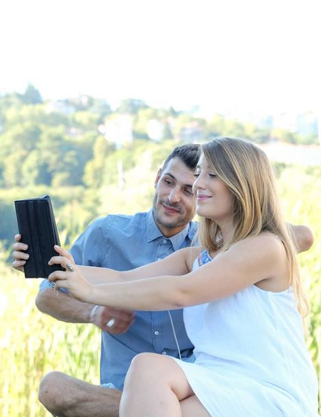 Gelukkig jong studenten paar met behulp van digitale tablet en mobiele telefoon met oordopjes buiten in de zomer seizoen met groene stad achtergrond - Foto, afbeelding